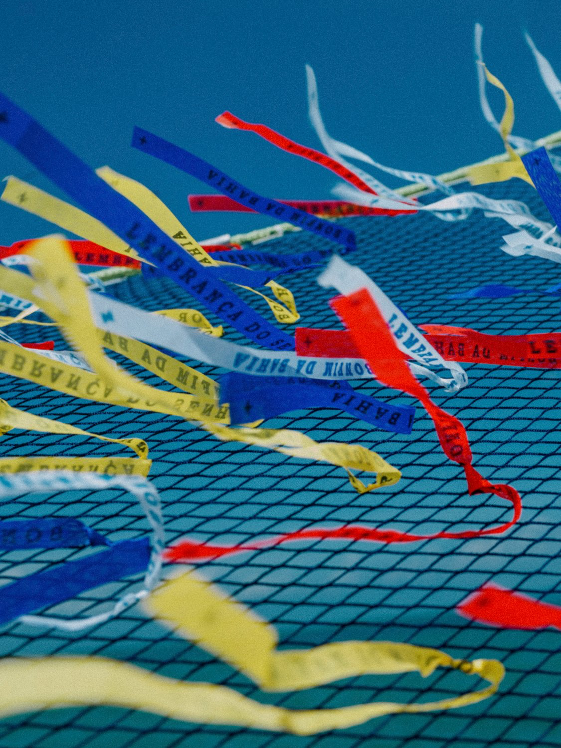 Multicoloured Senhor do Bonfim wish ribbons printed with 'Lembrança do Senhor do Bonfim da Bahia', caught in a net against a deep blue sky