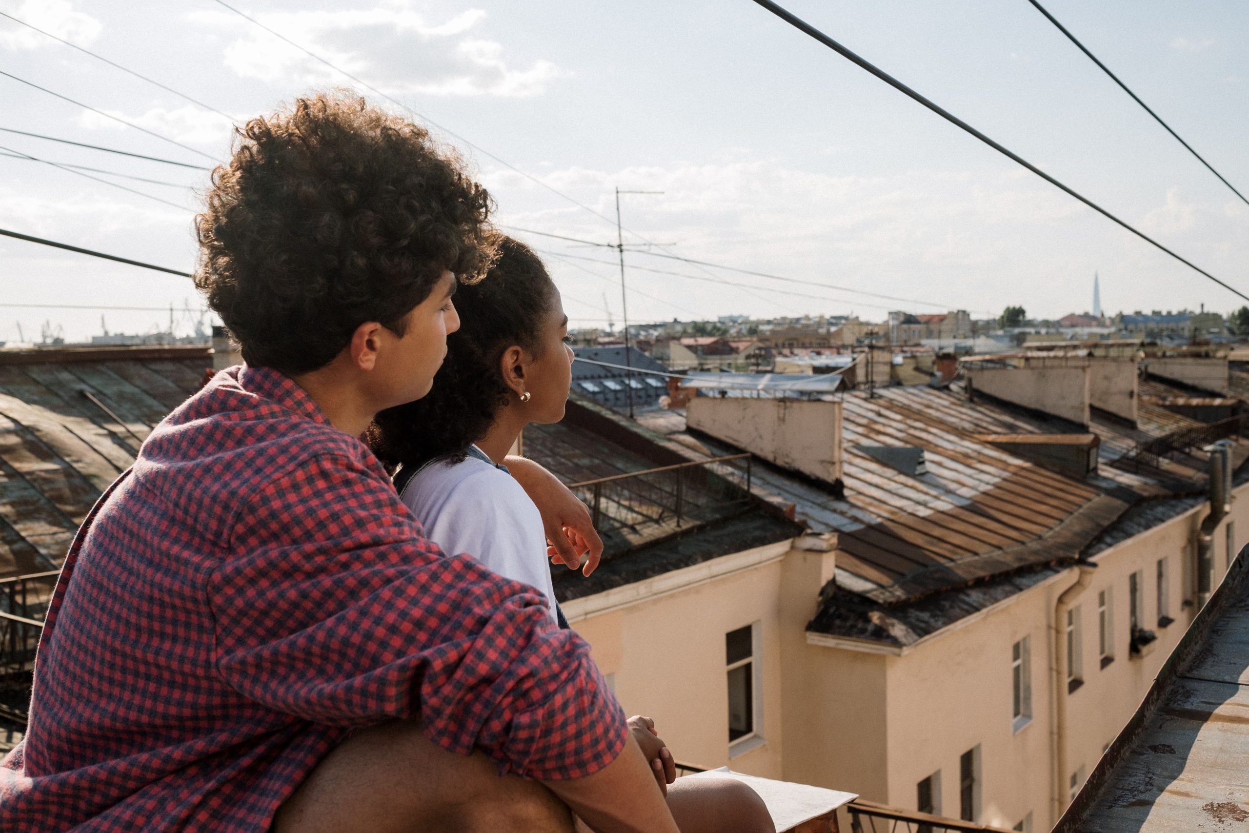 Two young people sit side by side on a rooftop railing, looking out over a city skyline